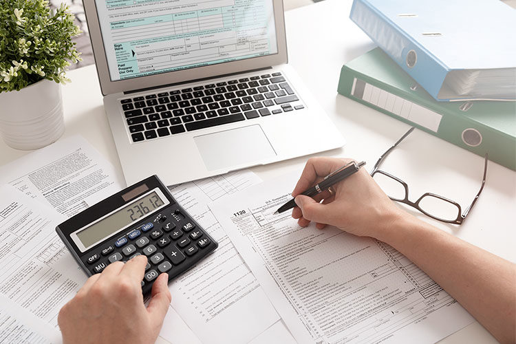 Imagem de uma pessoa escrevendo em um papel, com uma calculadora e um notebook sobre a mesa